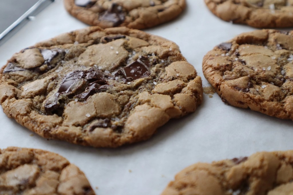 A close-up of freshly baked chocolate chip cookies with a golden brown color and sea salt on top, arranged on parchment paper.