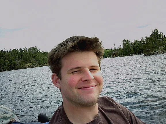 A young man with short brown hair smiles at the camera while sitting by a lake, surrounded by trees.